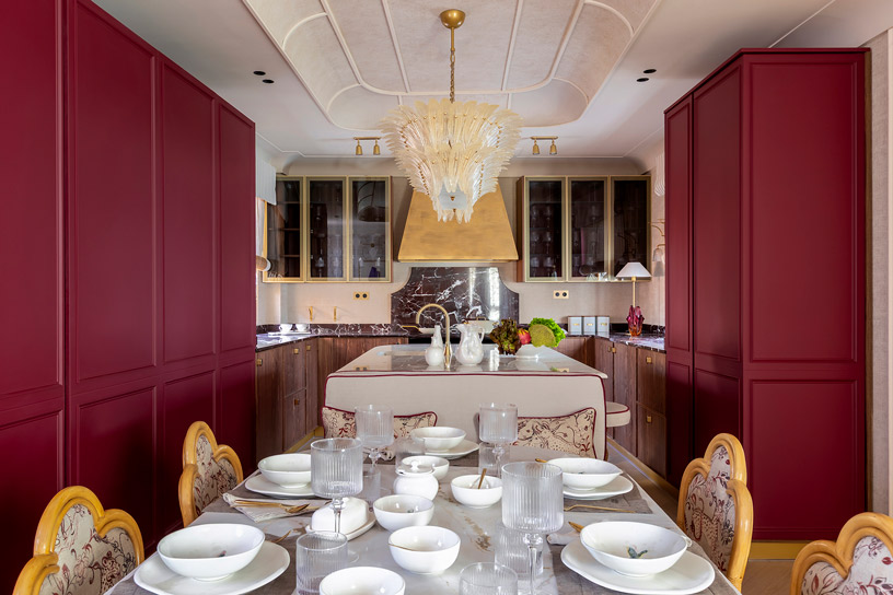 Cocina de diseño contemporáneo en Casa Decor 2026 con muebles en rojo lacado, gran isla central y lámpara escultórica, combinación de mármol, madera y detalles dorados en un espacio elegante y sofisticado.