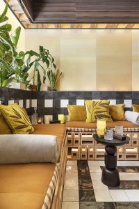 Salón con banco perimetral revestido en azulejo cerámico pequeño de Nais, cojines mostaza, suelo geométrico y plantas, diseño de Estudio Querencia en Casa Decor 2026