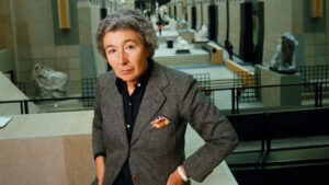 Retrato de Gae Aulenti, arquitecta y diseñadora italiana, con chaqueta gris y camisa negra, apoyada sobre una estructura de piedra en el interior de un museo, con esculturas, vitrinas y elementos arquitectónicos al fondo.