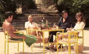 Escena de la película «La piscina» (1969), con cuatro personajes sentados alrededor de una mesa de jardín amarilla, acompañada de sillas de estructura tubular del mismo color, pertenecientes a la colección Locus Solus de Gae Aulenti.