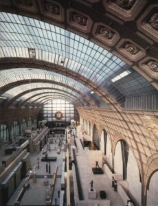Vista panorámica del interior del Musée d’Orsay de París, rehabilitado por Gae Aulenti, con la gran bóveda acristalada de hierro y vidrio de la antigua estación ferroviaria, galerías distribuidas en varios niveles, esculturas y pasarelas de piedra clara.