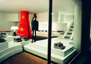 Interior del showroom de Olivetti en París, diseñado por Gae Aulenti, con máquinas de escribir y calculadoras expuestas sobre plataformas blancas escalonadas, una gran estructura roja y lámparas Pipistrello.