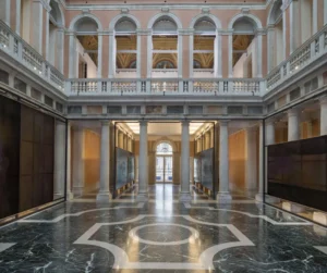 Patio interior del Palazzo Grassi de Venecia, rehabilitado por Gae Aulenti, con columnas y arcos de piedra, galerías superiores con balaustradas, paredes en tonos rosados y un suelo de mármol oscuro con motivos geométricos blancos.