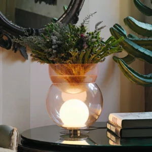Lámpara Giova de Gae Aulenti, diseñada en 1964, con base de cristal transparente, esfera luminosa blanca y recipiente superior de vidrio ámbar utilizado como jarrón con flores y ramas verdes, sobre una mesa auxiliar oscura.