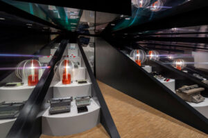Interior del showroom de Olivetti en Buenos Aires, diseñado por Gae Aulenti, con máquinas de escribir y calculadoras sobre plataformas blancas, superficies espejadas, estructuras oscuras y lámparas King Sun de tonos rojizos.
